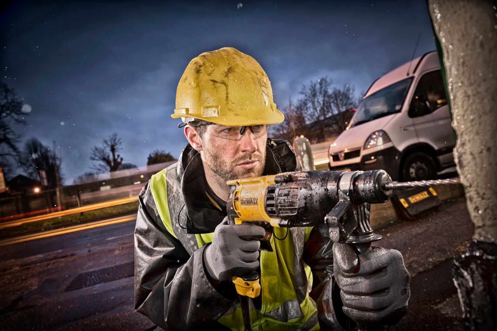 Un trabajador de la construcción con casco amarillo y chaleco reflectante utiliza un taladro percutor DEWALT al aire libre, con una furgoneta blanca y luces borrosas al fondo.