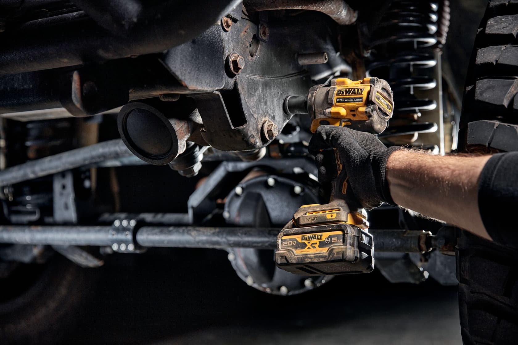 A person wearing gloves uses a yellow and black DEWALT cordless impact wrench (DCF922B model) to work on bolts under a vehicle. The tool is battery powered and designed for tough automotive tasks.