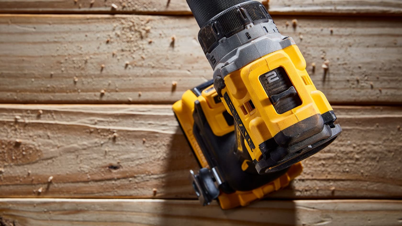 Primer plano de un taladro inalámbrico DEWALT siendo usado sobre una superficie de madera, con aserrín disperso alrededor. El taladro es amarillo y negro y está en funcionamiento.
