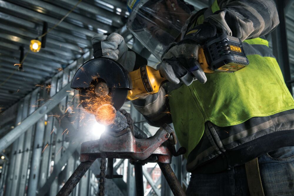 Un trabajador utiliza una amoladora angular inalámbrica DEWALT (modelo DCG414T2) para cortar metal, lo que genera chispas. El trabajador lleva equipo de seguridad, incluidos guantes, visera y chaleco reflectante, en un entorno industrial.