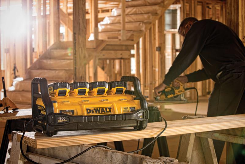 Una estación de energía portátil DEWALT con cuatro ranuras para baterías está ubicada sobre un banco de trabajo en un sitio de construcción. Una persona al fondo utiliza una herramienta eléctrica sobre una tabla de madera.