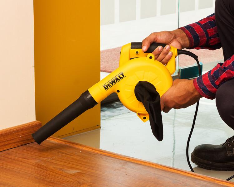 Una persona utiliza un soplador eléctrico amarillo DEWALT para limpiar el polvo de un suelo de madera junto a una pared.