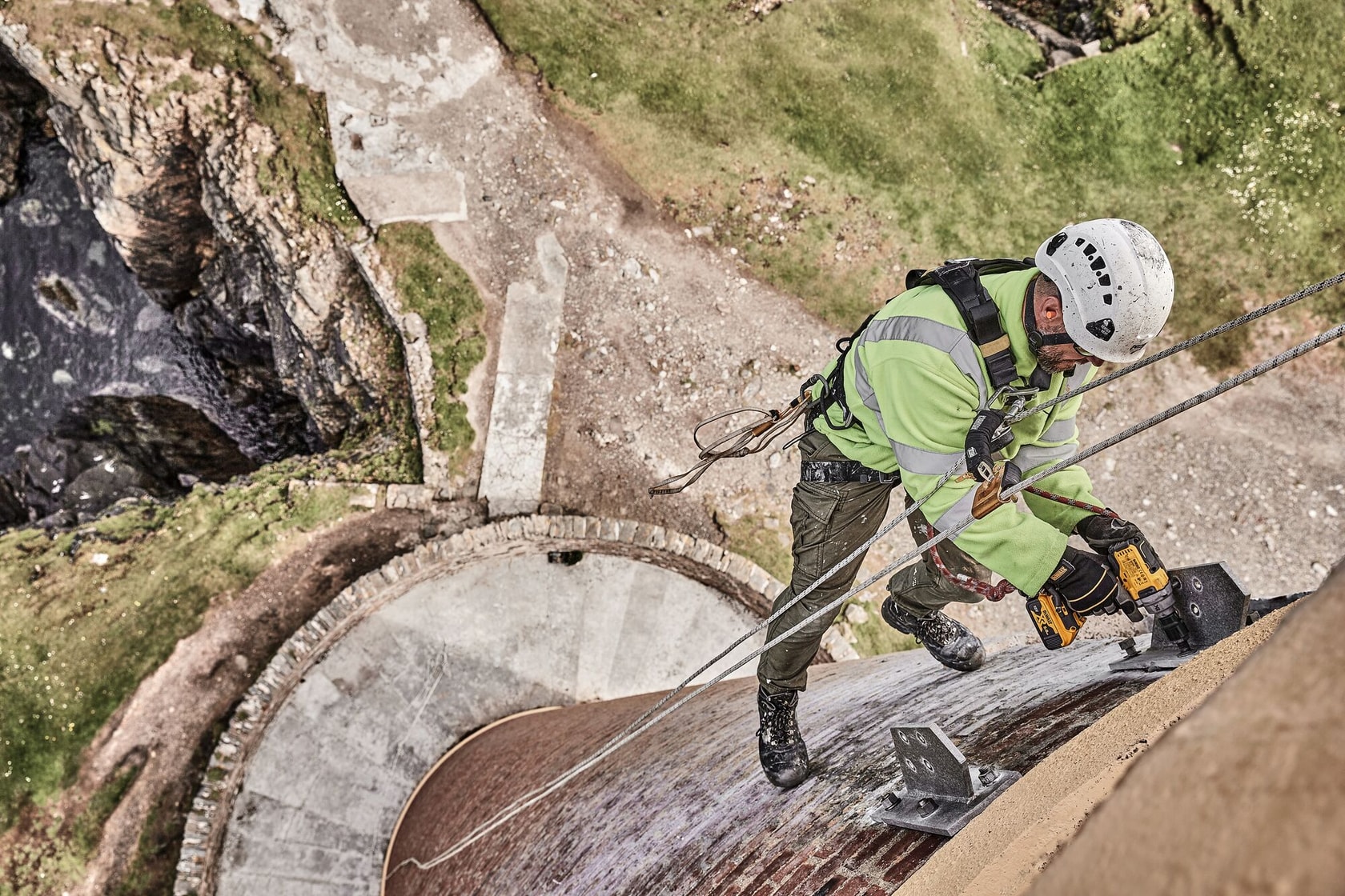 Trabajador usa herramienta DEWALT en escalada, reparando estructura exterior sobre piedra y césped.