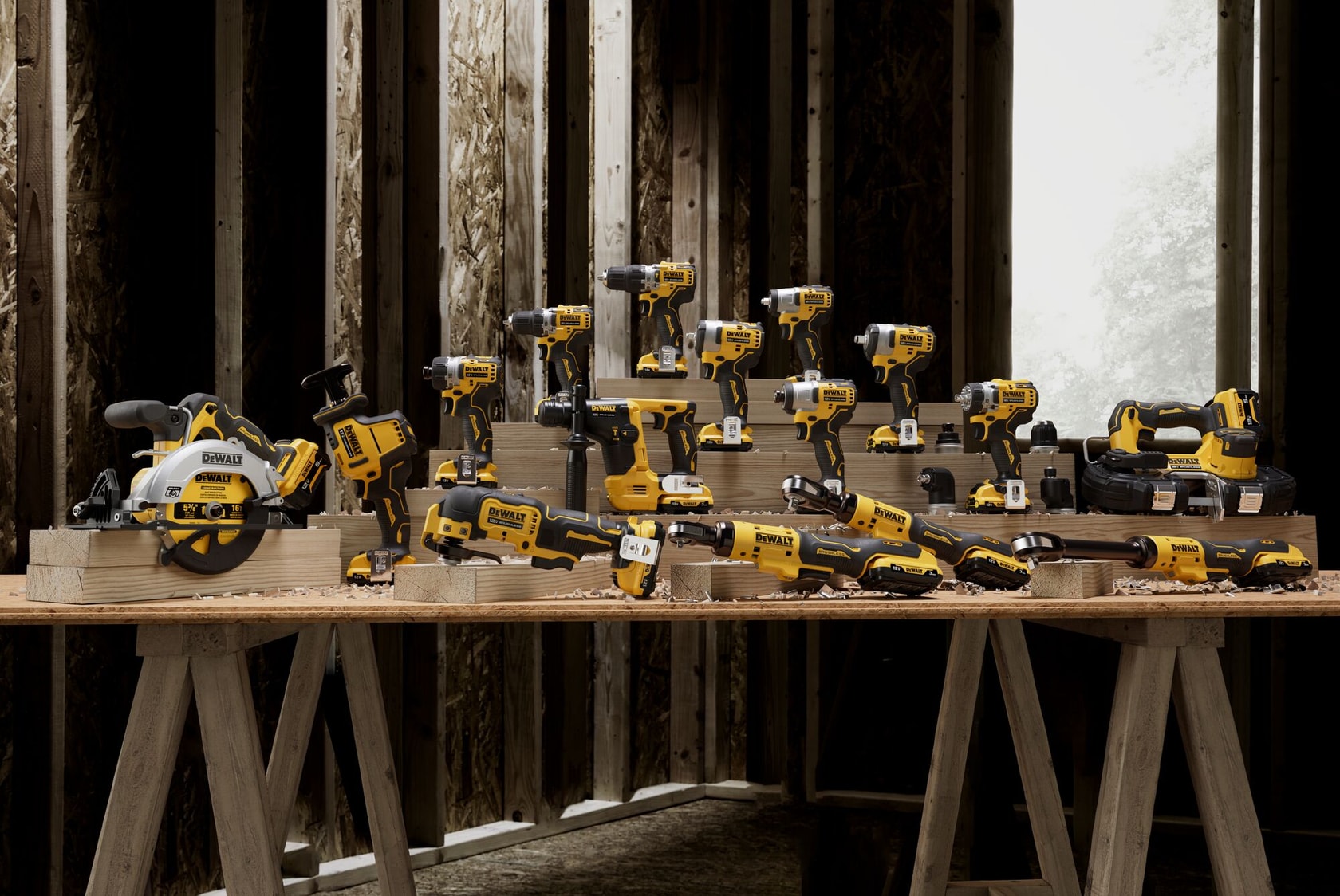 Una variedad de herramientas eléctricas inalámbricas DEWALT de color amarillo y negro, incluyendo taladros, sierras y destornilladores, están dispuestas sobre un banco de trabajo de madera en una habitación en construcción con marco de madera.