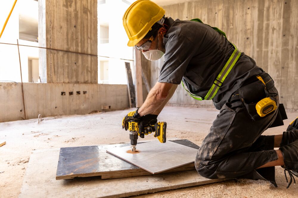 Trabajador usa taladro DEWALT en baldosa en obra, con equipo de seguridad y arrodillado.