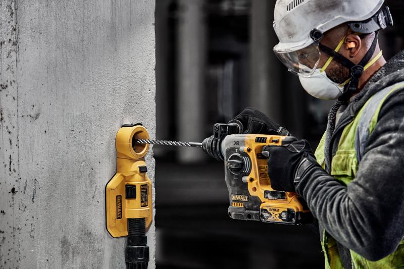 Un trabajador de la construcción con equipo de seguridad está utilizando un taladro eléctrico amarillo DEWALT para perforar una pared de concreto. Un accesorio de extracción de polvo DEWALT está conectado a la pared para recoger los residuos.