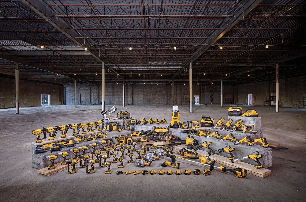 Una amplia selección de herramientas eléctricas, baterías y accesorios DEWALT en color amarillo y negro están exhibidos sobre bloques de concreto y tablas de madera, organizados dentro de un gran almacén vacío con techos y pilares expuestos.