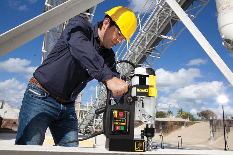 Una persona con casco amarillo y camisa azul opera un taladro magnético DEWALT DWE1622K sobre una superficie metálica en un sitio industrial al aire libre.
