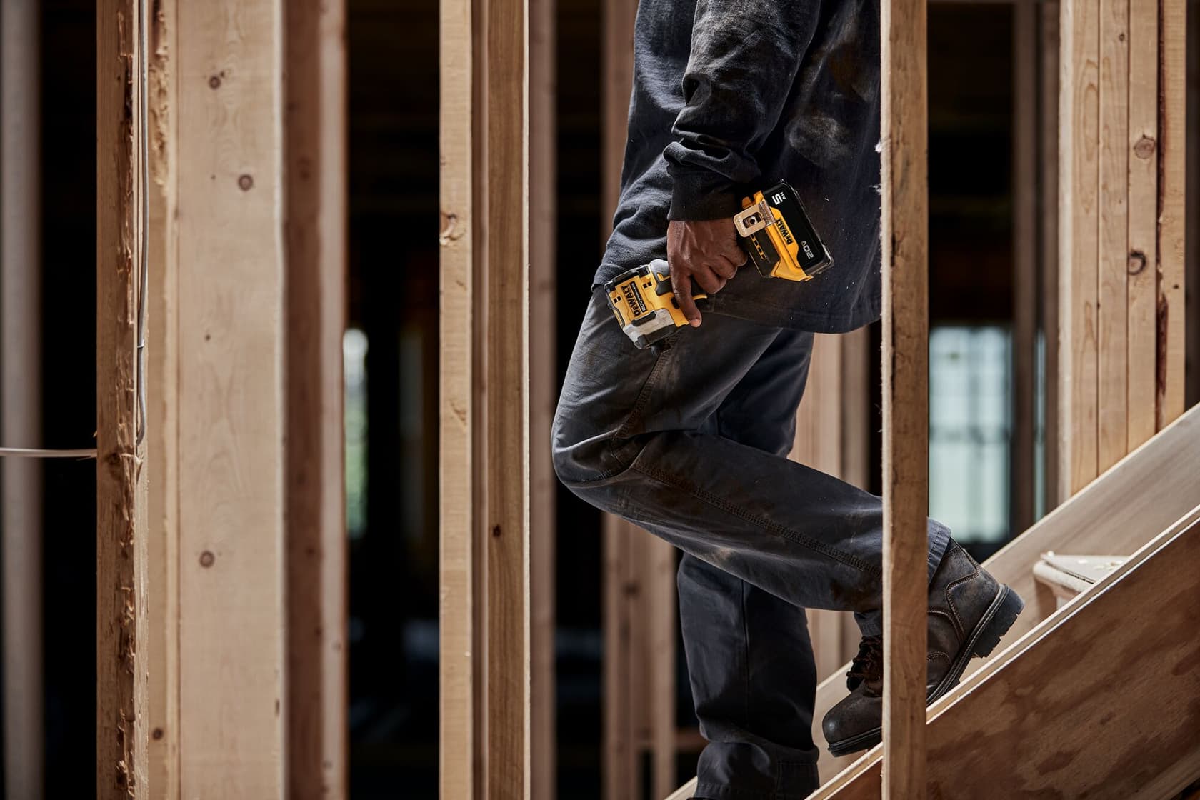 Una persona vestida con ropa de trabajo sube una escalera de madera en un edificio en construcción, sosteniendo una herramienta eléctrica inalámbrica amarilla y negra DEWALT.