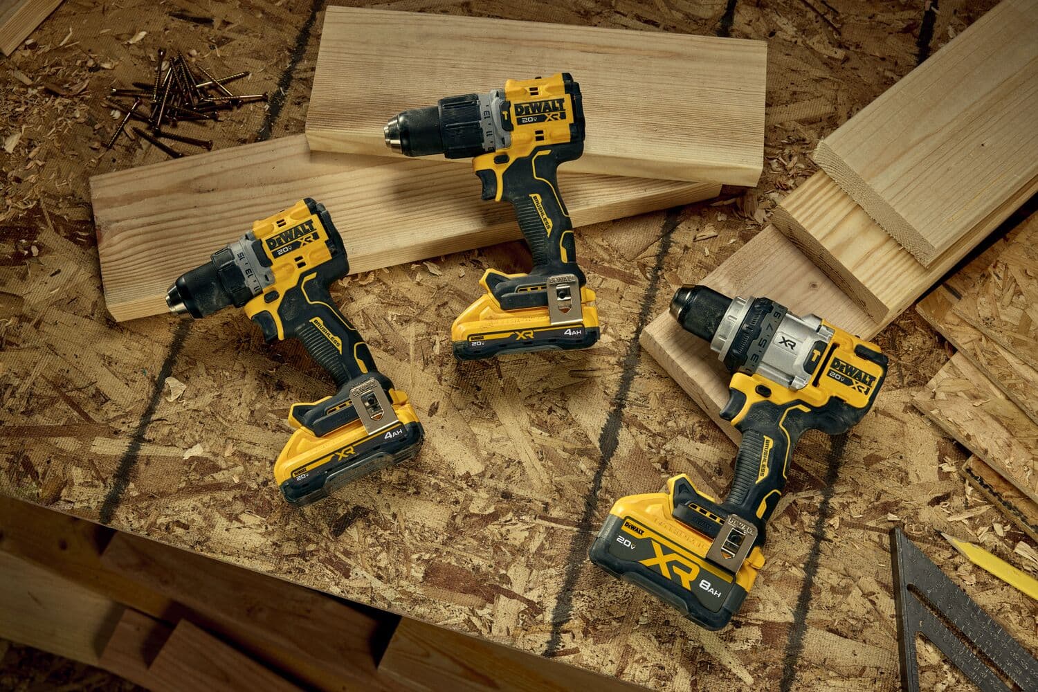 Tres taladros eléctricos inalámbricos DEWALT están colocados sobre una mesa de trabajo de madera junto a tablas de madera y clavos. Los taladros son de color amarillo y negro, y algunos tienen etiquetas 'XR' en sus baterías, indicando mayor duración. La escena sugiere un entorno de taller.